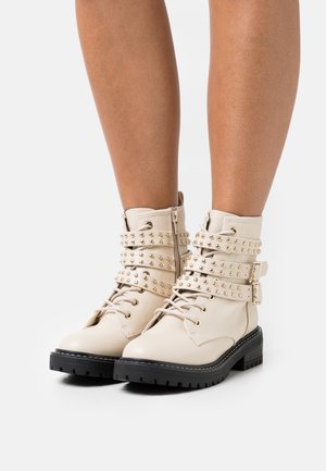 Beige veganskt läder stövletter med spikade remmar, snörning och en klumpig svart gummisula. Har dekorativa metalldetaljer.