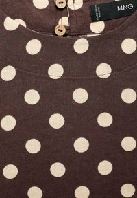 Hnědá tkanina s velkými krémovými puntíky, s kulatým výstřihem, zapínáním na knoflík na zadní straně a štítkem s pokyny k údržbě viditelným na límci.
