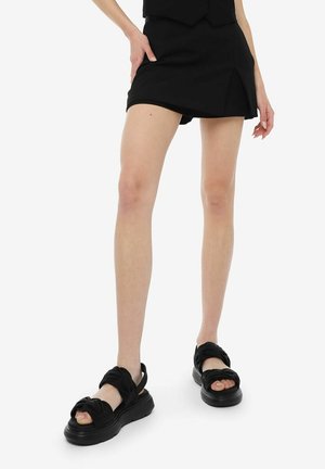 Femme portant une mini-jupe noire et des sandales à plateforme noires avec des brides torsadées, debout sur fond blanc.