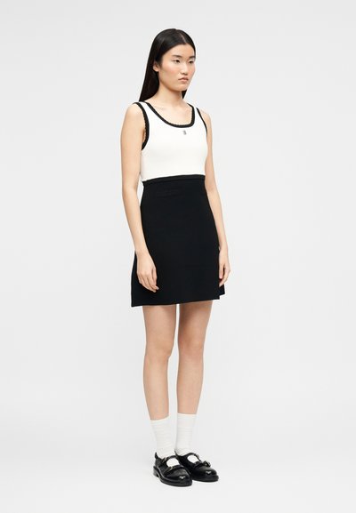 Γυναίκα όρθια που φοράει λευκό τοπ χωρίς μανίκια με μαύρη διακόσμηση, μαύρη φούστα, λευκές κάλτσες και μαύρα δερμάτινα παπούτσια.