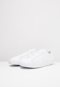 Zapatillas blancas con parte superior de cuero liso, puntera redonda y suela plana de goma. Presenta ojales y detalles cosidos a lo largo del costado.