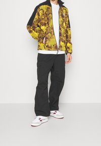 Giacca con zip a motivi gialli e marroni, maniche nere, abbinata a pantaloni cargo neri e sneaker bianche, impreziosita da dettagli color bordeaux.