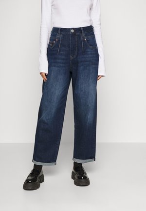 Høj talje mørkeblå denim jeans med løs pasform, med rullemanchetter, forlommer og synlige syninger. Båret med skinnende sorte loafers.