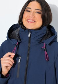 Usmívající se žena upravuje šňůrky kapuce na tmavě modré mikině na zip s fialovými šňůrkami a zipovou kapsou na prsou na bílém pozadí.