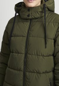 Olijfgroene pufferjack met hoge kraag, ritssluiting en zwarte trekkoorden. Opvallend door de gewatteerde, padded textuur.