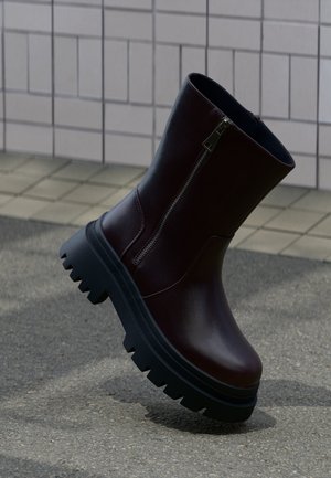 Botte en cuir marron foncé avec une épaisse semelle noire à crampons et une fermeture éclair latérale, posée sur un sol carrelé et asphalté.