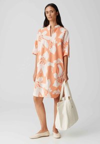 Korte mouwen, perzikkleurige jurk met grote witte bloemenprint. Heeft een kraagloze halslijn. Het model draagt een witte tote tas en beige ballerina's.