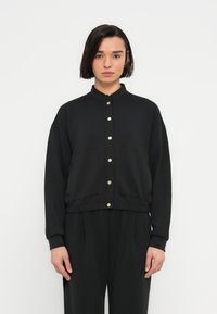 Γυναίκα με κοντά σκούρα μαλλιά, ντυμένη με μαύρο σακάκι με χρυσά κουμπιά και μαύρο παντελόνι που ταιριάζει, στέκεται μπροστά σε ένα απλό λευκό φόντο.