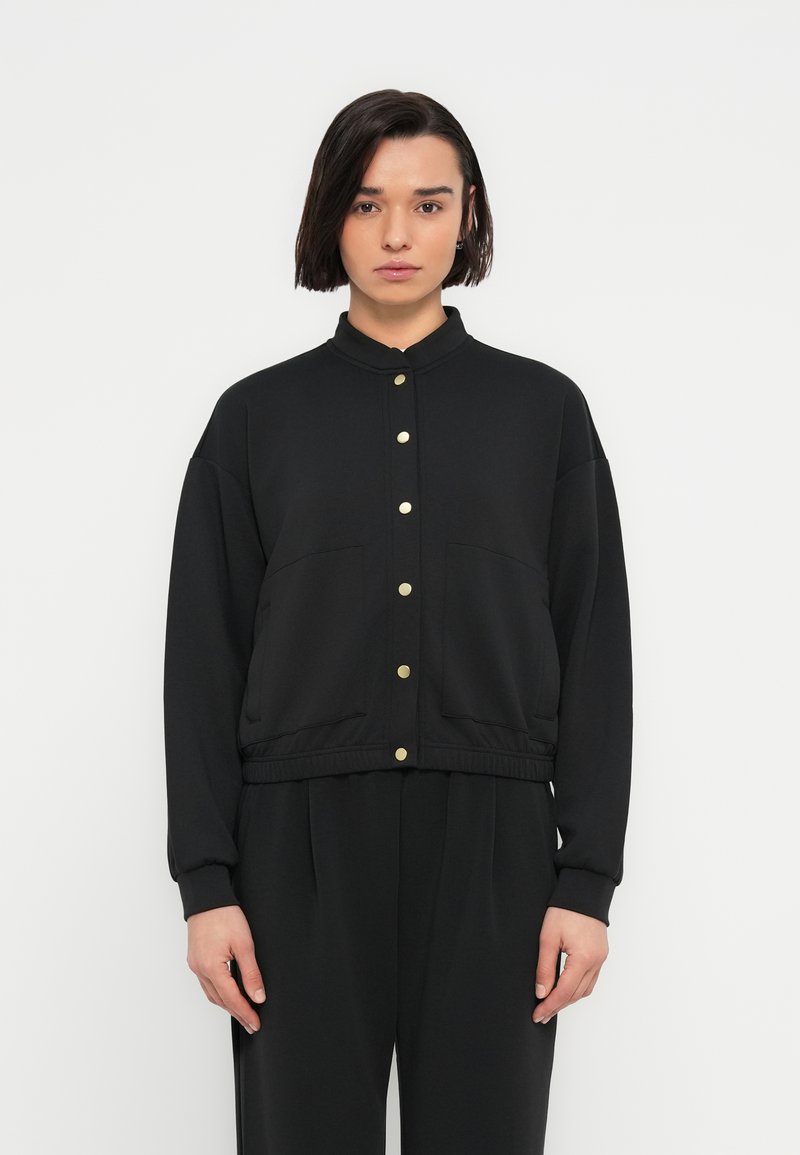 Γυναίκα με κοντά σκούρα μαλλιά, ντυμένη με μαύρο σακάκι με χρυσά κουμπιά και μαύρο παντελόνι που ταιριάζει, στέκεται μπροστά σε ένα απλό λευκό φόντο.