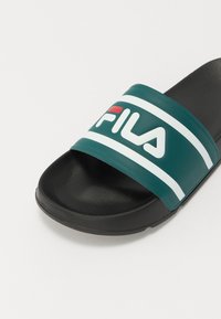 Žalios ir juodos šlepetės su plačiu diržu, kuriame yra baltos juostelės ir "Fila" logotipas baltos ir raudonos spalvos. Textūruota padas.