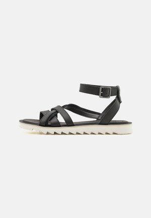 Sandale en cuir noir avec des brides entrecroisées, boucle à la cheville et semelle blanche crantée. Design plat, minimaliste avec une texture lisse.
