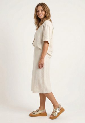 Frau steht seitlich, trägt ein beiges Leinenkleid und goldene Plateausandalen aus Metall, lächelt vor einem einfarbigen hellen Hintergrund.