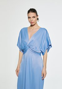 Svijetloplava haljina od svilenog materijala s dubokim V-izrezom, zavezana u struku, kratkih rukava i lepršavim siluetom.