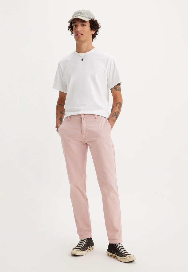 Pantaloni in cotone rosa con tasche laterali, abbinati a una maglietta bianca. Il modello indossa un cappellino grigio chiaro e sneakers nere.