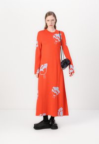 Lungo vestito rosso a maglia con motivo floreale blu, scollatura rotonda e maniche lunghe. Abbinato a una borsa rotonda nera e scarpe nere chunky.