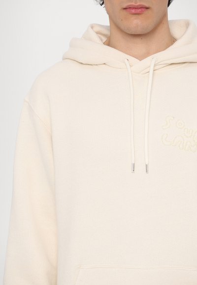 Cremfarbener Hoodie aus strukturiertem Stoff, mit Känguru-Tasche, Zugbändern und gesticktem "Soul Land"-Logo auf der Vorderseite.