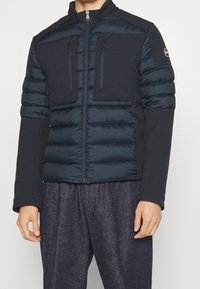 Marinblå puffjacka med en åtsittande design, som har två zippered bröstfickor, släta tygsektioner och en hög krage.