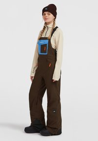 Bruine geïsoleerde overall met een blauwe borstzak en oranje ritsaccenten, gedragen over een crèmekleurig langetermijnshirt, in combinatie met zwarte laarzen.