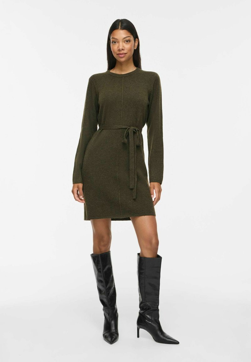 Femme portant une robe olive verte mi-longue à manches longues avec ceinture à la taille et bottes noires à talons montant aux genoux, debout sur un fond uni.