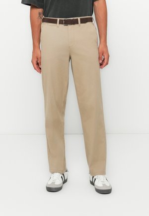 Light beige chinos with a straight leg fit, featuring a brown belt. Worn with white and gray sneakers displaying black accents.