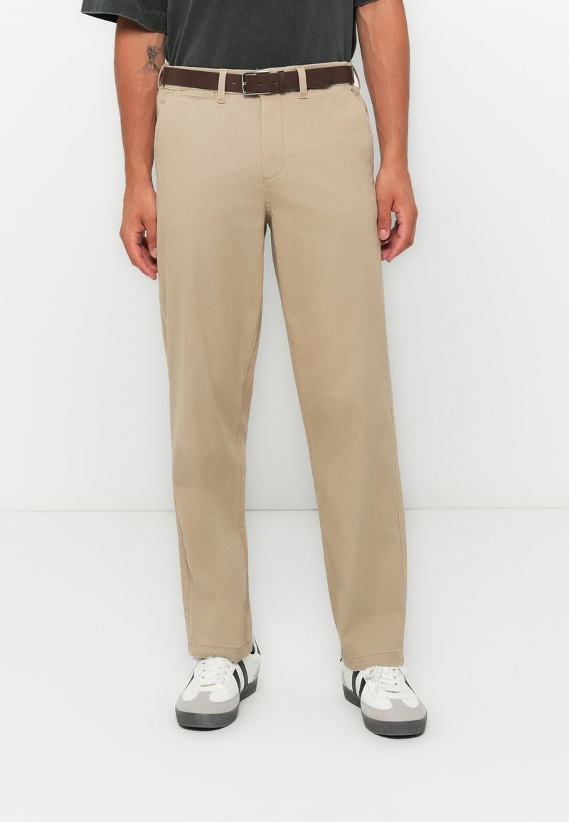 Light beige chinos with a straight leg fit, featuring a brown belt. Worn with white and gray sneakers displaying black accents.