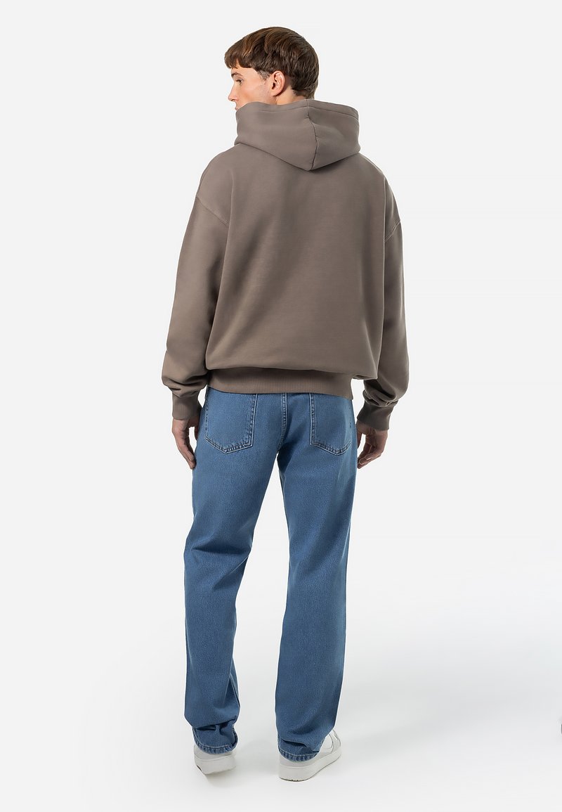 Bruine hoodie, ontspannen pasvorm, ribgebreide manchetten en onderrand. Gecombineerd met lichtblauwe rechte spijkerbroek en witte sneakers.