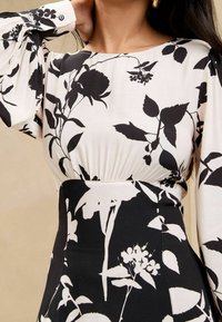 Kobieta w dopasowanej czarno-białej sukience z długim rękawem i kontrastowym kwiatowym wzorem, z uniesioną ręką do szyi.