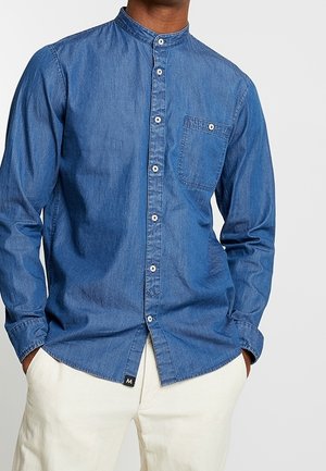 Homme portant une chemise en jean bleue à boutons avec col mao et un pantalon blanc, les mains dans les poches, sur un fond uni.