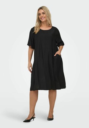 Kvinne iført en løs, knelang svart kjole med korte ermer, en hånd i lommen, smilende, stående mot en ensfarget bakgrunn.