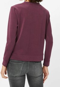 Sweatshirt bordeaux à manches longues et col rond, doté d'une texture lisse et d'un ourlet légèrement raccourci. Porté avec un jean gris.
