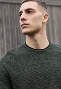 Vihreä neulottu neulepaita, jossa on teksturoitu kuvio ja musta pyöreä kaulus. Sileä kangas, vartalonmyötäinen malli ja hienovaraisia yksityiskohtia.