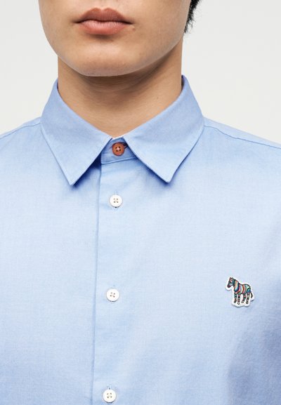 Άτομο που φοράει ανοιχτό μπλε πουκάμισο με κουμπιά και μια μικρή πολύχρωμη κηφισιανή ραφή στη αριστερή πλευρά του στήθους.