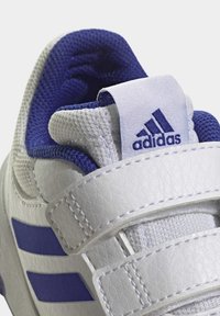 Zapatilla blanca con material de cuero, acentos morados, un diseño texturizado y correas de Velcro ajustables. Logo de Adidas en el talón.