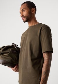 Bărbat cu braț tatuat, îmbrăcat cu un tricou verde măsliniu, ținând o geantă de voiaj de culoare verde închis și maro, pe un fundal deschis simplu.
