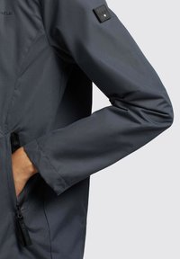 Dunkelgraue, wasserdichte Jacke mit glatter Textur, ausgestattet mit einer Reißverschlusstasche und einem Logopatch am Ärmel. Figurbetonte Passform.