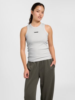 Vrouw met lang donker haar in een paardenstaart, gekleed in een lichtgrijs mouwloos geribbeld topje en donkergrijze losse broek, staand met één hand in de zak.