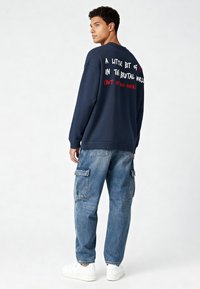 Jūras spēku kreklis ar baltu un sarkanu tekstu aizmugurē, apvienots ar gaiši ziliem cargo džinsiem un baltiem kedām. Kokvilnas materiāls, brīvs griezums.