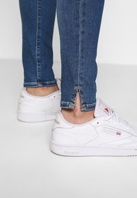Witte Reebok sneakers in combinatie met donkerblauwe skinny jeans. De jeans hebben een splitzoomdetail bij de enkels. De schoenen hebben een gestructureerd leer en een klassiek ontwerp.