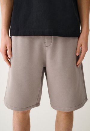 Primo piano di una persona che indossa pantaloncini beige larghi e una camicia nera con le mani appoggiate ai fianchi su uno sfondo uniforme.