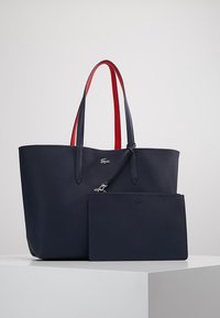 Sac cabas bleu marine avec poignées bordées de rouge et pochette détachable assortie, tous deux ornés de petits logos crocodile, posés sur une surface blanche.