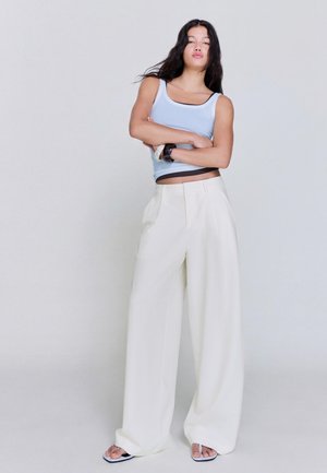 Mladá žena s dlhými tmavými vlasmi stojí so založenými rukami, oblečená v svetlomodré tielko, vysoký pás široké biele nohavice a sandále.