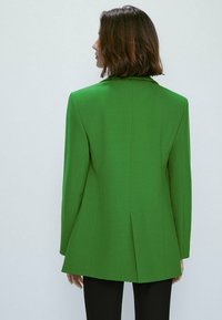 Zielona marynarka dopasowana o strukturalnym kształcie, ostre klapy i jedno rozcięcie z tyłu. Połączona z czarnymi spodniami, gładka faktura materiału.