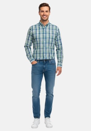 Hombre sonriente de pie con una mano en el bolsillo, vistiendo una camisa a cuadros verde y azul abotonada, vaqueros azules y zapatillas blancas sobre fondo blanco.