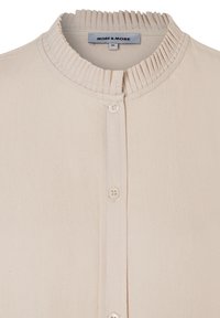 Beige Hemd mit Knopfleiste und geripptem Kragen, das über eine glatte Textur verfügt und drei Knöpfe auf der Vorderseite hat. Markenetikett sichtbar.