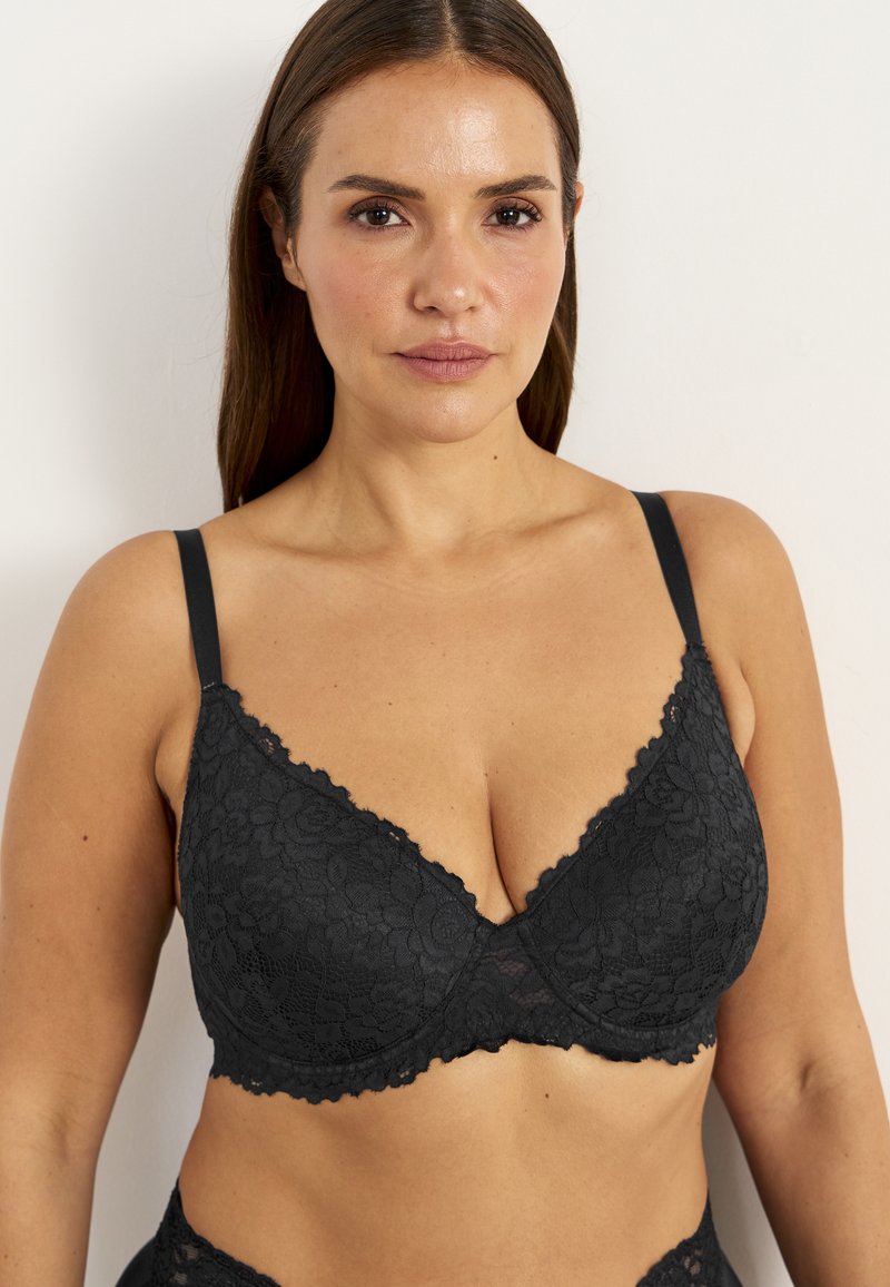 Musta pitsinen rintaliivi, jossa on kukkakuvio, tukeva langanveto, säädettävät hihnat ja aaltoilevat reunat. Pehmeä pinta syvällä V-muotoisella designilla.