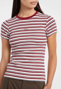 Donna che indossa una maglietta a maniche corte in maglia a costine con strisce orizzontali rosse, bianche e blu navy e il logo "Guess Jeans" sul petto.
