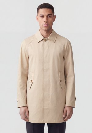 Uomo che indossa un cappotto beige monopetto con tasche con patta abbottonate e cinturini ai polsini su sfondo semplice grigio chiaro.