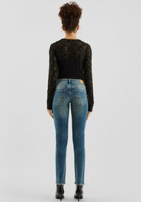 Czarna koronkowa bluzka z długim rękawem, dopasowane niebieskie dżinsy z lekkim gradientem oraz czarne botki na obcasie. Widoczne tylne kieszenie na dżinsach.