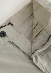 Zbliżenie na pas beżowych spodni, pokazujący guzik, częściowo rozpięty metalowy suwak oraz szczegółowe szwy.