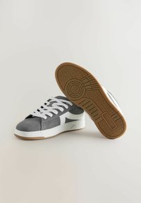 Baskets en suède gris avec des accents blancs, présentant une semelle blanche plate et une semelle extérieure en caoutchouc brun texturé. Des lacets blancs épais complètent le design.